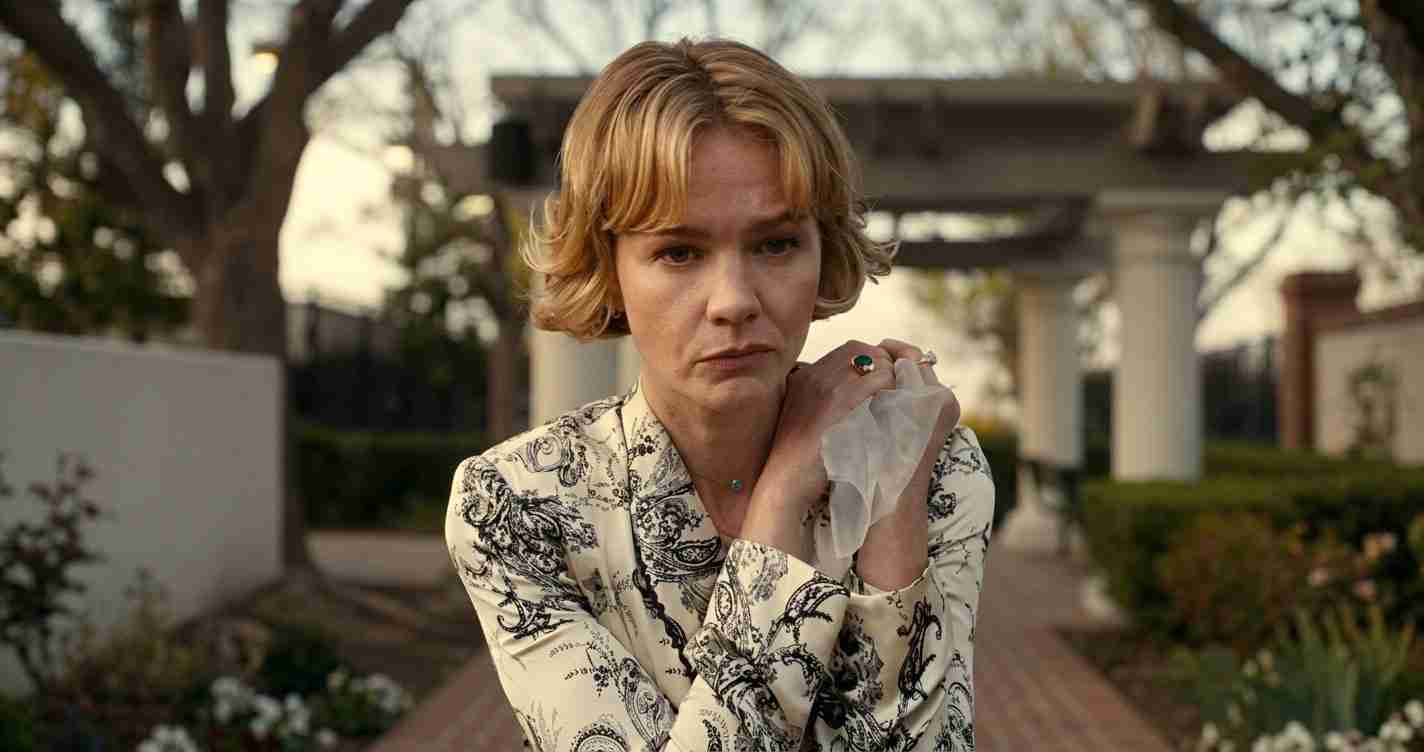 Woman with short blonde hair in a paisley-patterned jacket looks worried, standing in a garden courtyard.