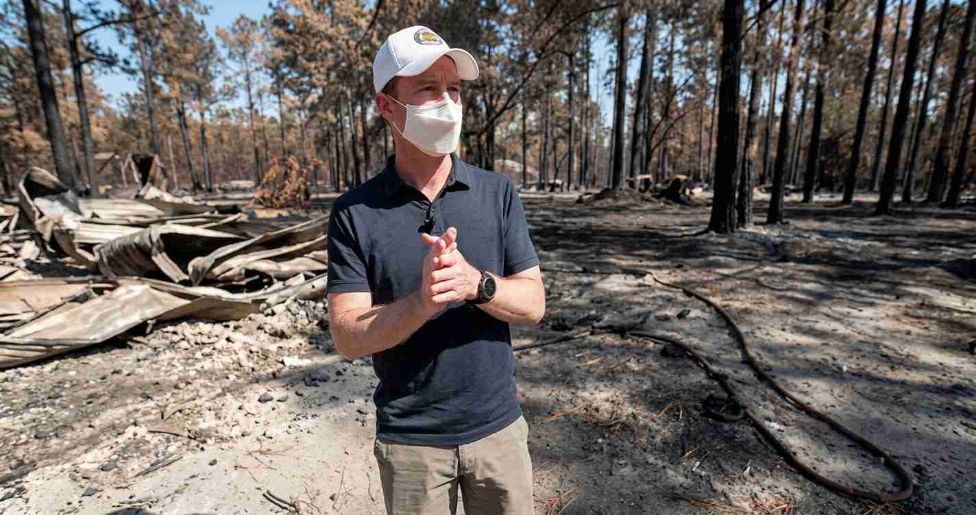 Man wearing a mask and cap stands in a burned forest with charred trees and debris, hands clasped.