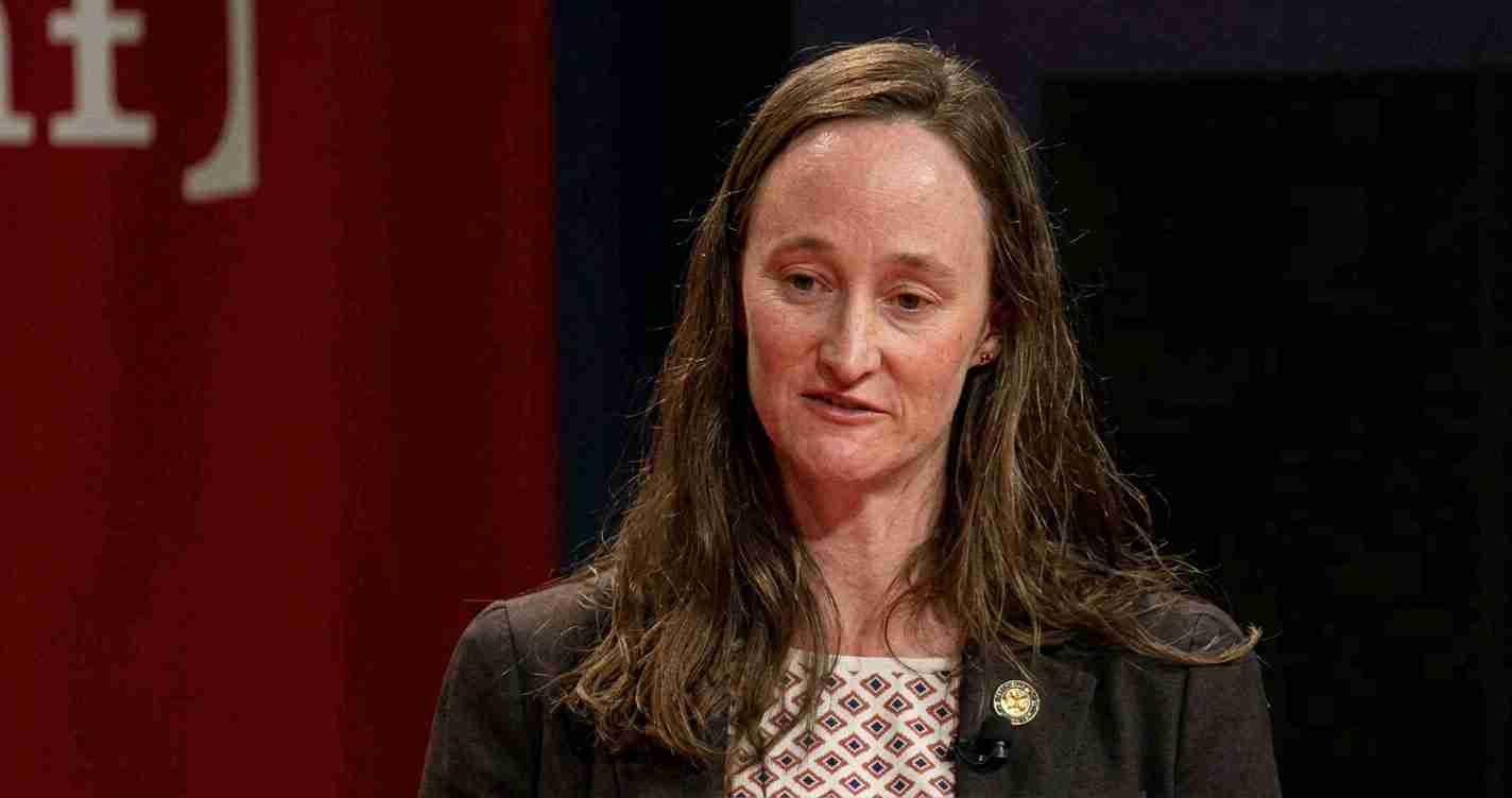 Woman in a blazer speaking on stage, wearing a geometric patterned top and a round pin on her lapel.