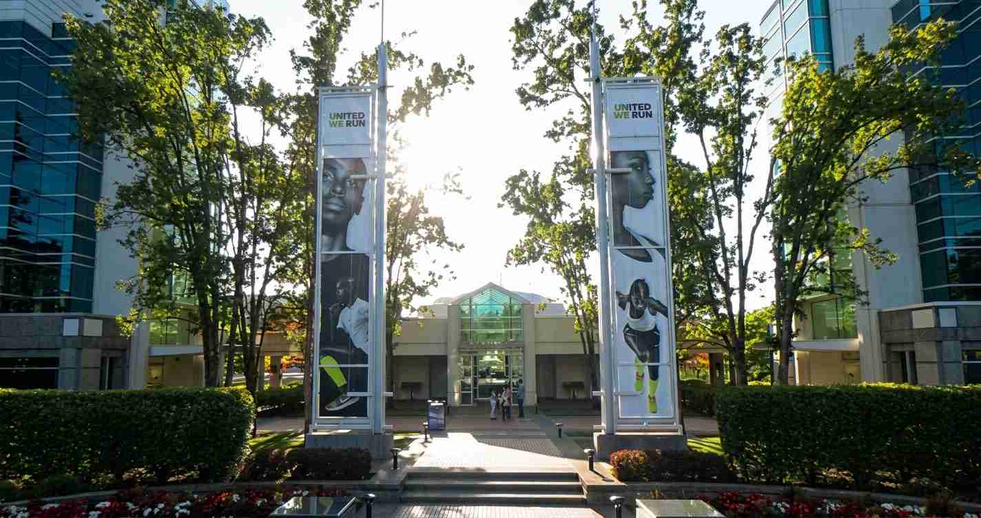 Two tall banners reading UNITED WE RUN flank a glass entrance to a modern building, with trees and a sunny courtyard area.