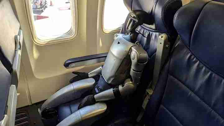 Silver humanoid robot seated in an airplane seat by the window, with arms folded and legs bent, as if traveling as a passenger.