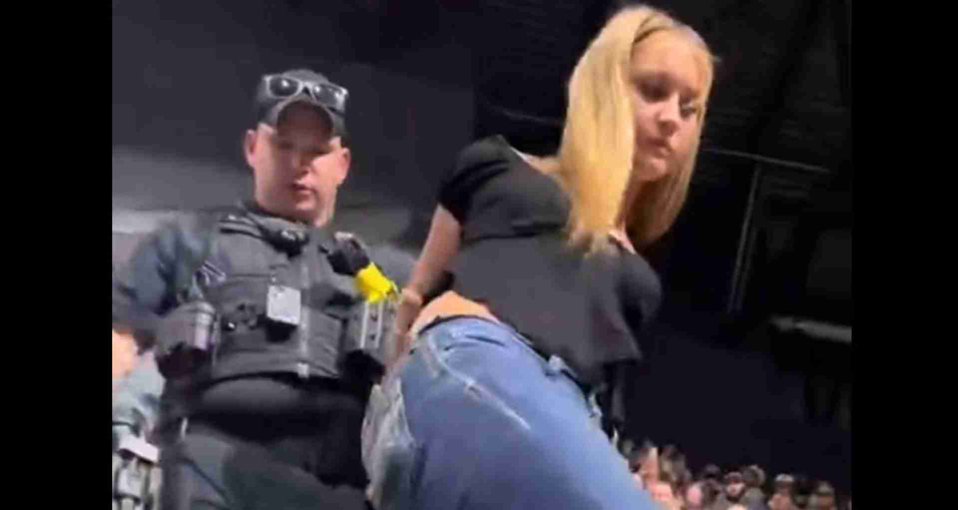 Security guard in tactical vest and cap standing on stage beside a blonde woman who is bending forward, with a crowd visible in the background.