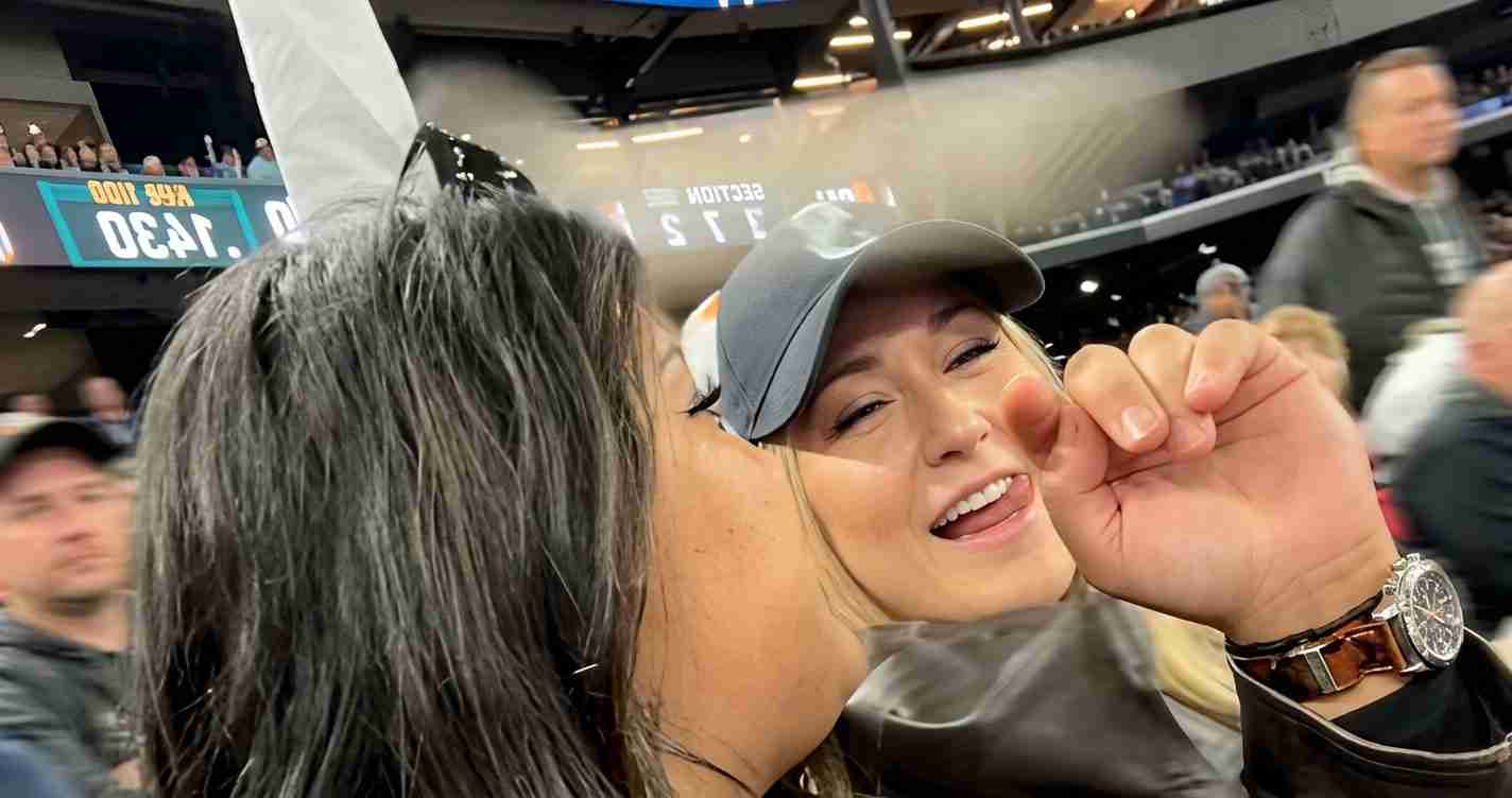 Two people in a crowded stadium share a kiss on the cheek; one wears a gray cap and a watch visible on the wrist.