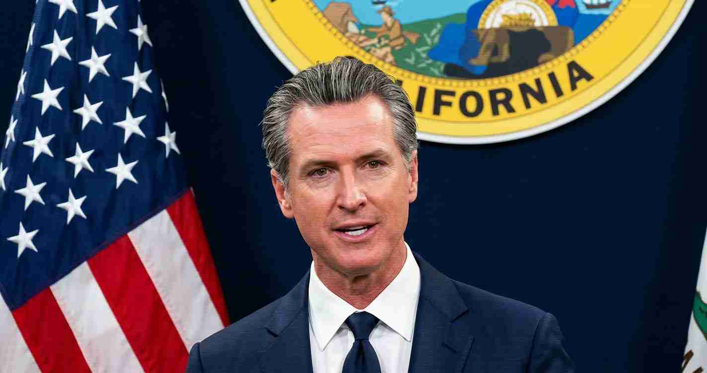 Male official in a navy suit delivers a speech at a press briefing, with the U.S. flag and California state seal in the background.