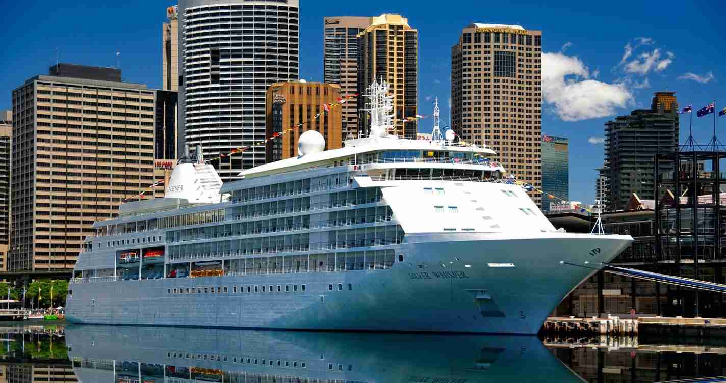 Large white cruise ship docked at a city harbor with tall office buildings in the background.