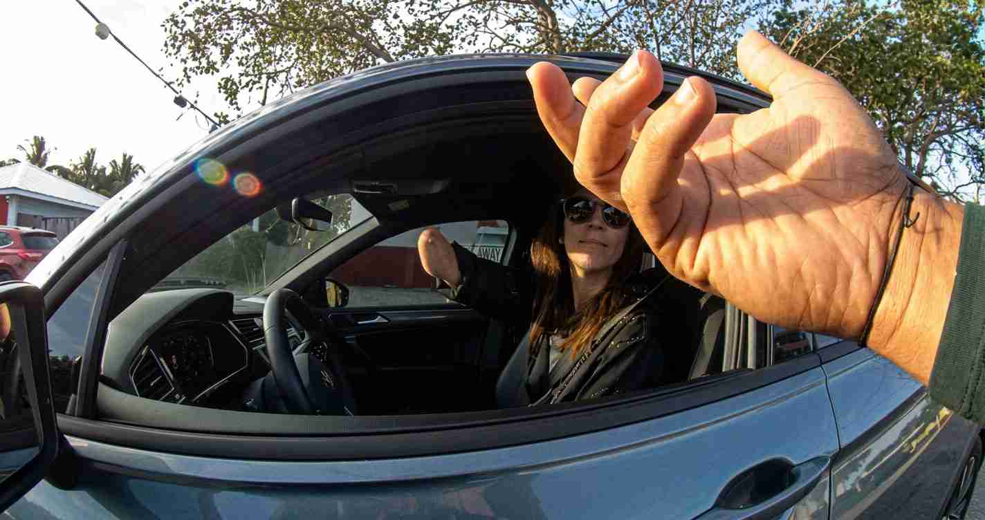 Woman in sunglasses seated in a blue car, arm extended toward the camera from the open window.