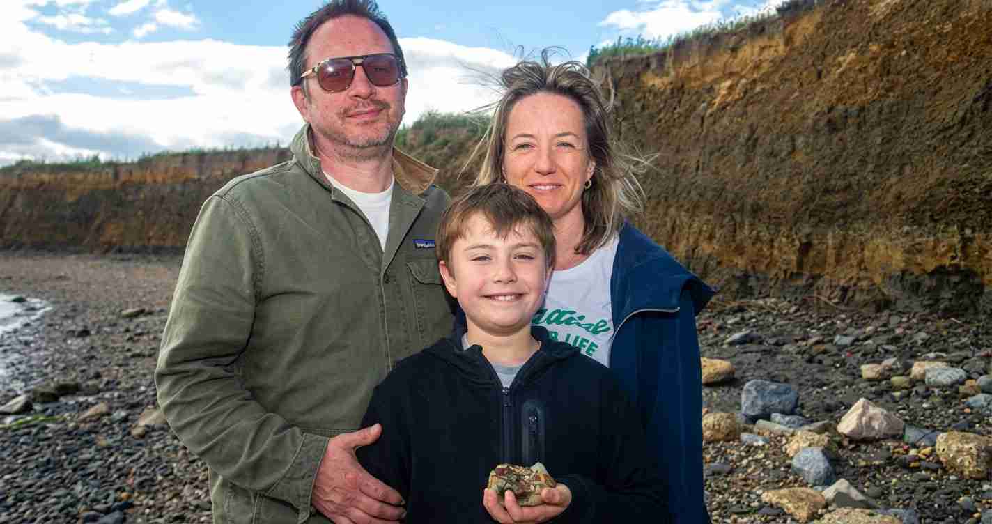Family of three posing together on a rocky shoreline, with a cliff in the background; man in sunglasses and green jacket, woman in a blue coat, and a smiling boy holding a rock.