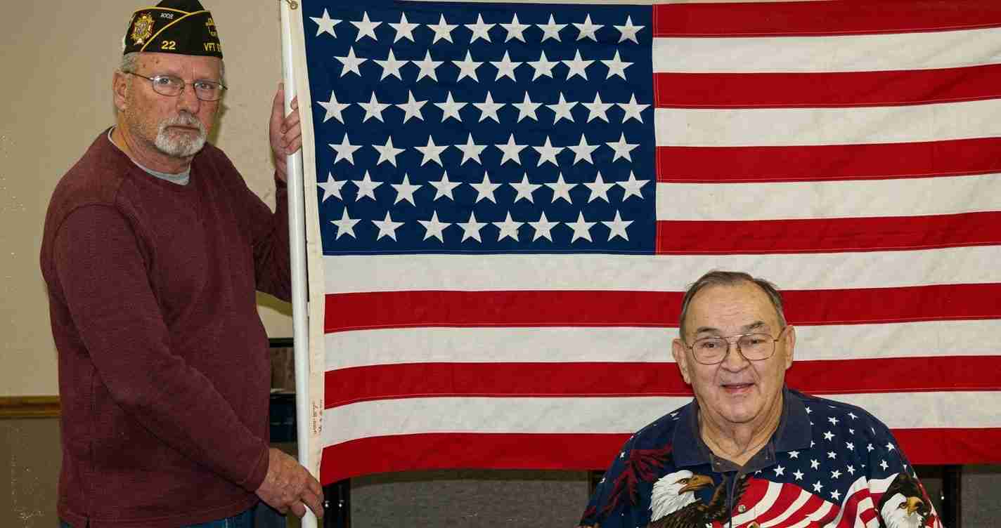 Two veterans pose beside a large American flag, one standing in a maroon sweater wearing a veterans cap and the other seated in a patriotic shirt.