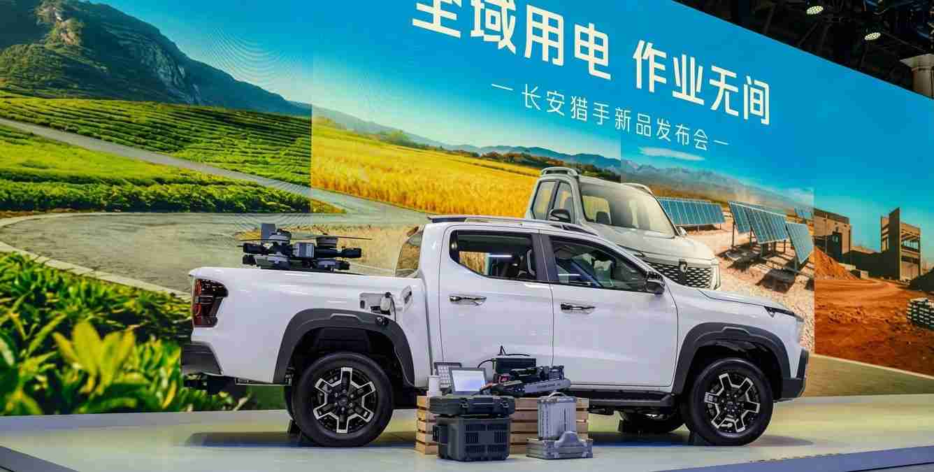 White pickup truck on an exhibition stage with gear in the bed and stacked boxes nearby; blue backdrop with Chinese text.
