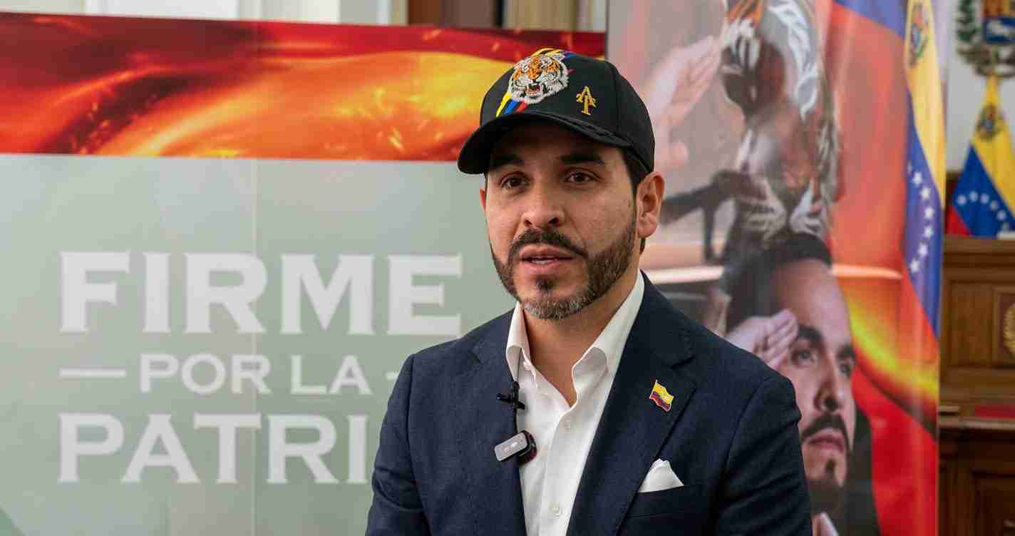 Man in a dark blazer and white shirt wearing a cap with a tiger emblem, speaking in front of banners.