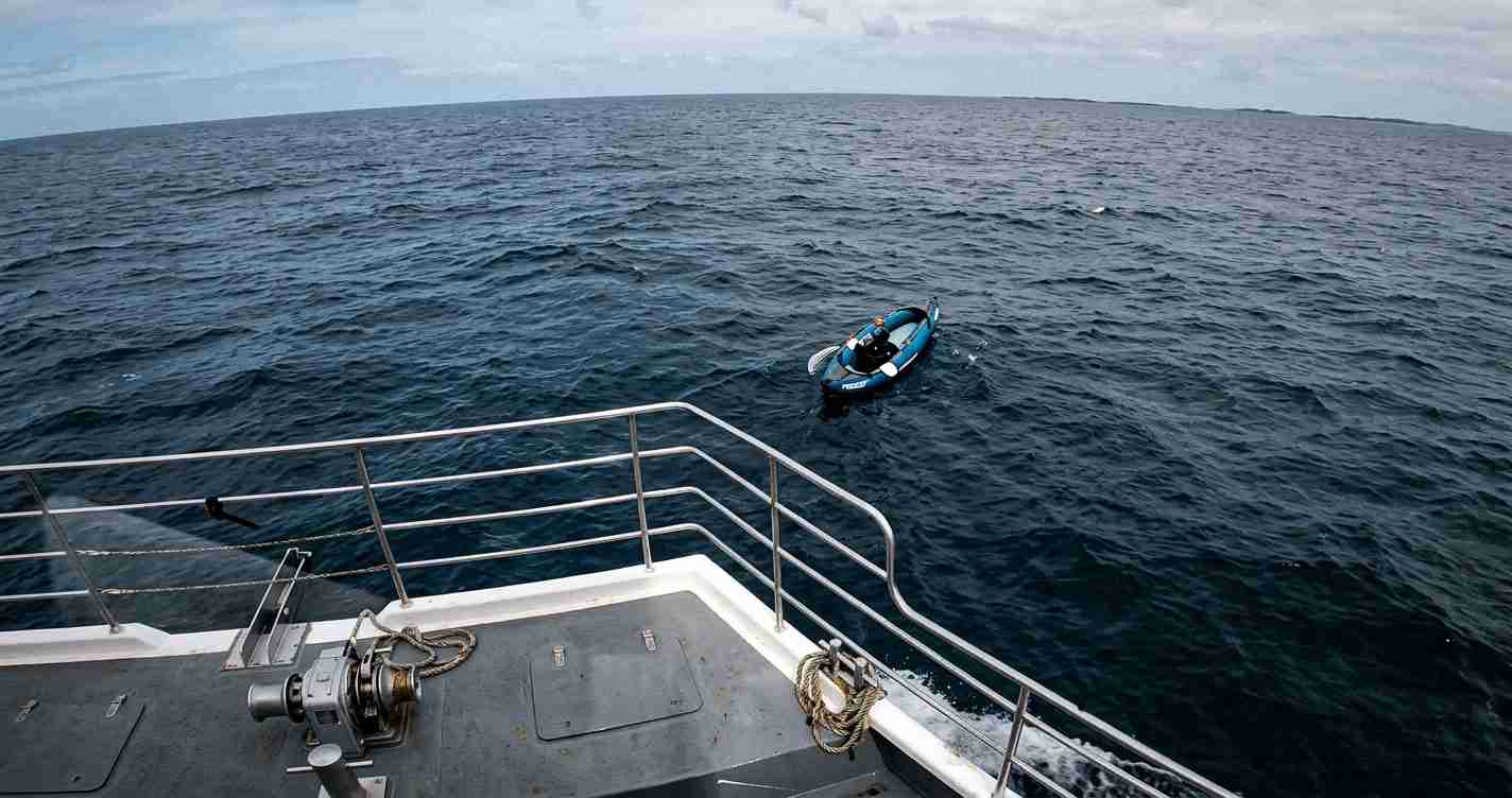 Small blue inflatable dinghy beside a larger ship's deck, floating on the open ocean under a cloudy sky.