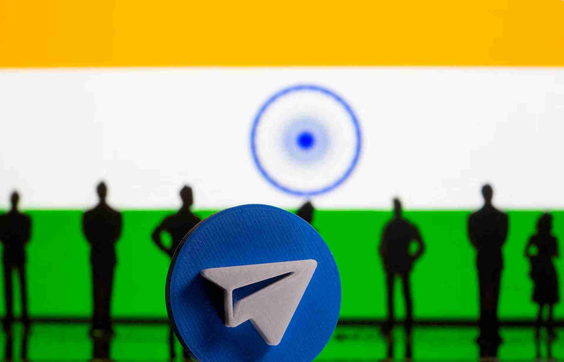 Blue circular token with a white angular logo in the foreground; silhouettes of people stand on a green field with a white-and-blue background and a large circular motif in the distance, suggesting a crypto/tech presentation.