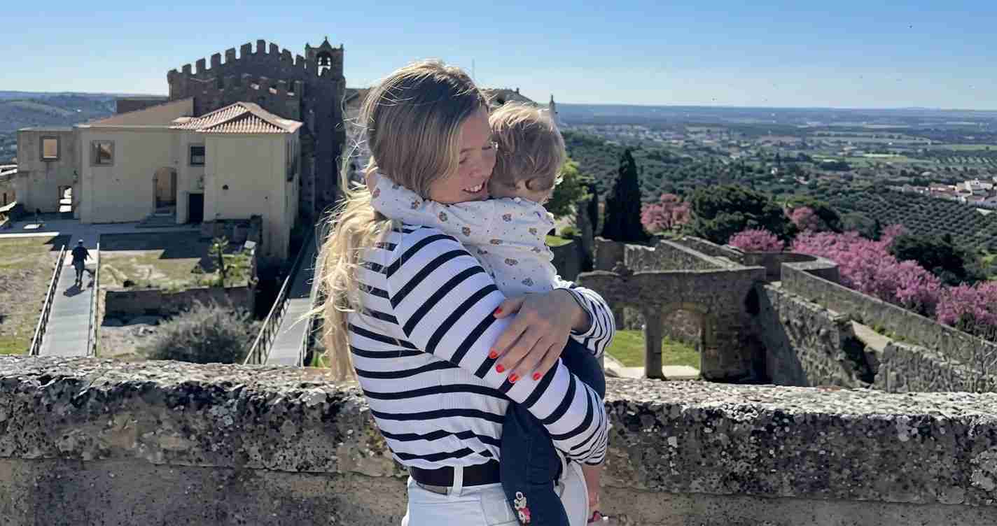 Woman in a striped shirt hugging a child on a stone fortress wall, with a historic castle and green valley in the background.