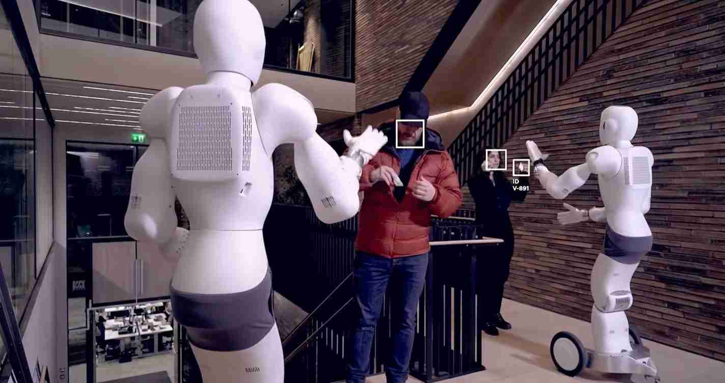 Two white humanoid robots shake hands with a man in a red jacket during a robotics demonstration in a modern building lobby.