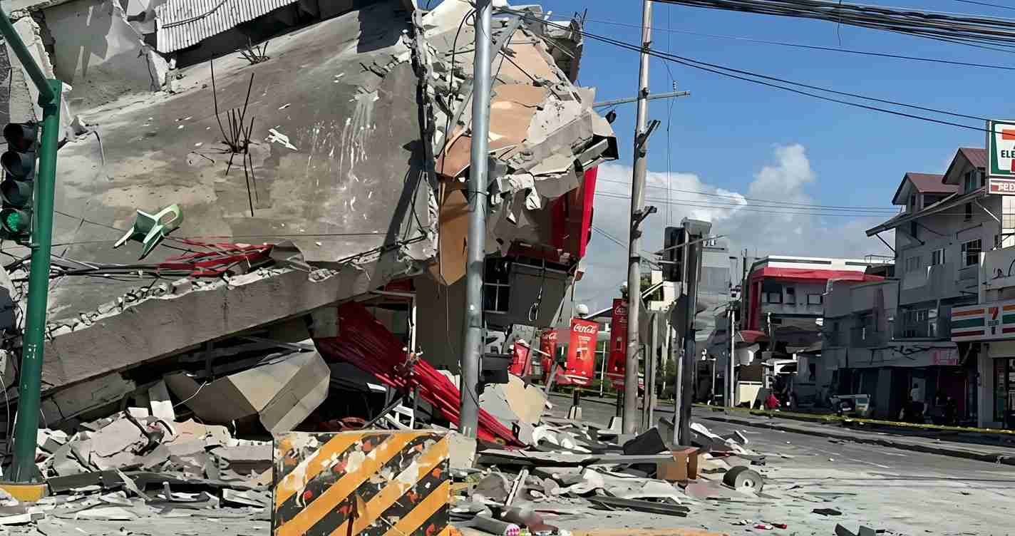 Rubble and a collapsed building clog a street, with bent utility poles and scattered debris in the foreground.
