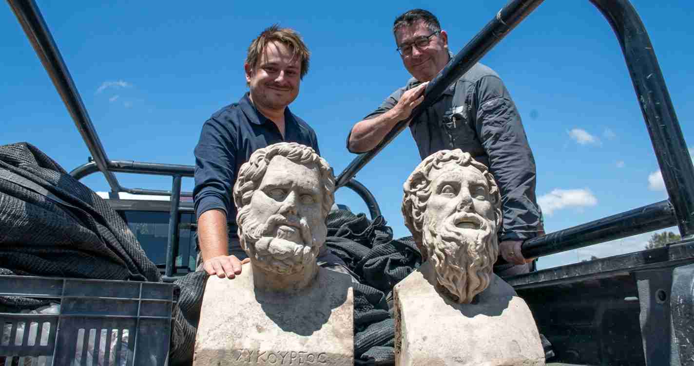 Two workers stand in a truck bed beside marble busts of bearded men against a blue sky.