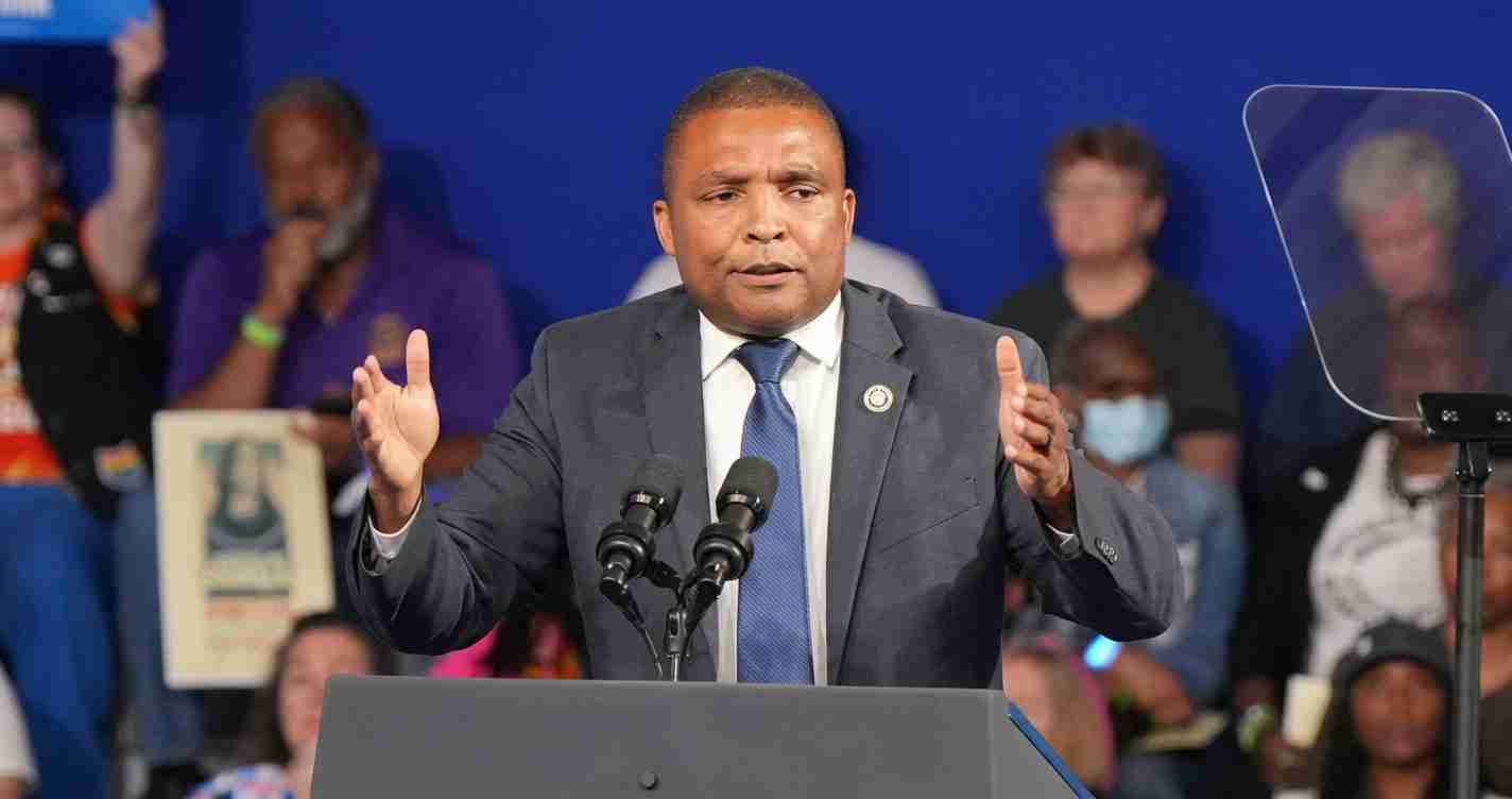 Man in a suit speaks at a podium with two microphones, gesturing with his hands in front of a blue backdrop behind a crowd of listeners.