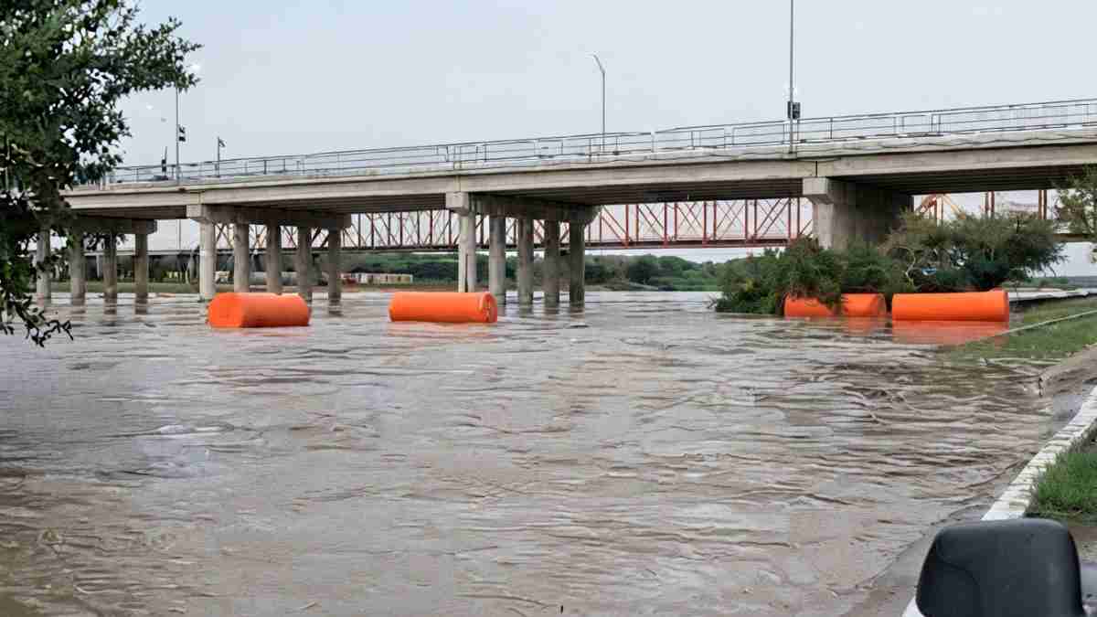 Flooded river under a bridge with orange floating barriers and submerged trees nearby.