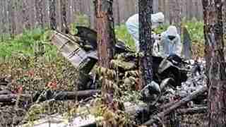 Two people in white hazmat suits examine a crashed, rusted aircraft fuselage in a dense forest.