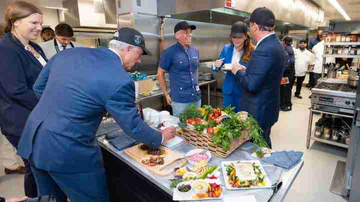 Group of professionals in a stainless-steel kitchen sampling plated dishes laid out on a counter with a basket of vegetables nearby.