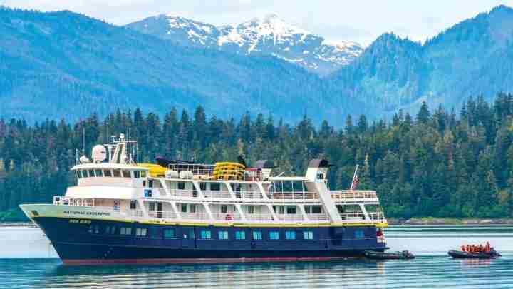Large blue and white tour yacht on calm water with forested mountains in the background.