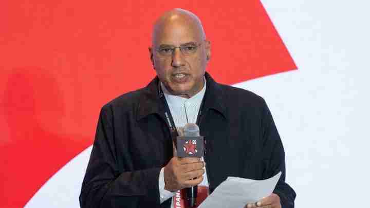 Man in a dark jacket speaks at a conference, holding a microphone and notes against a red backdrop behind him.
