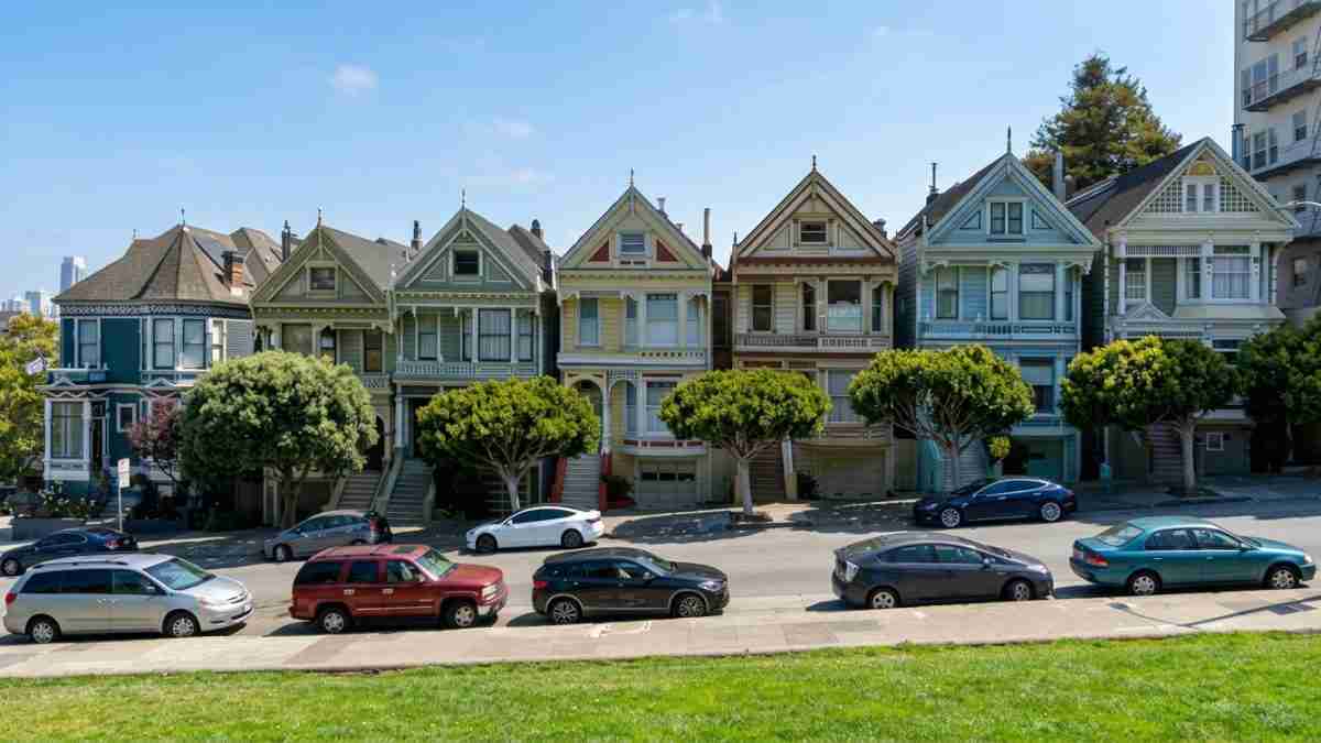 Row of colorful Victorian houses along a street with parked cars and green trees under a blue sky.