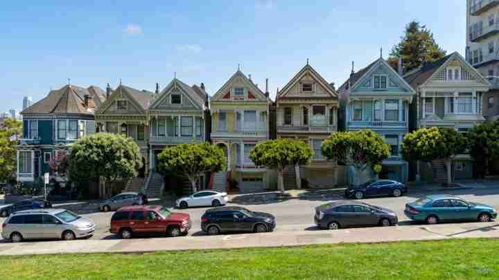 Row of colorful Victorian houses along a street with parked cars and green trees under a blue sky.