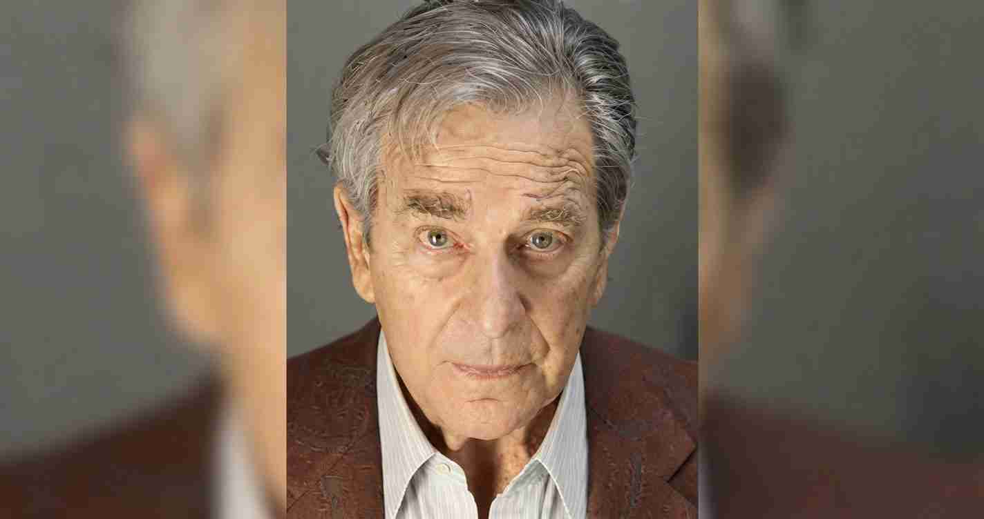 Close-up portrait of an older man with silver hair and a thoughtful expression, wearing a brown blazer and light shirt.