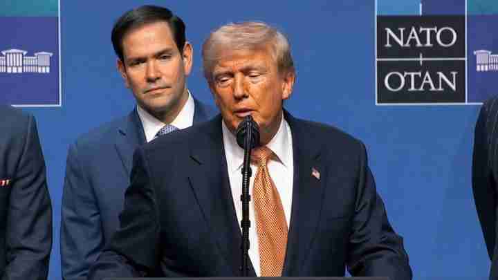 Donald Trump speaking at a NATO press conference, with a NATO sign visible in the background