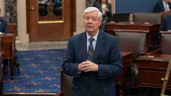 Adult man in a dark suit and striped tie stands in a wood- and marble-lined legislative chamber, hands clasped as he speaks to lawmakers or an audience.