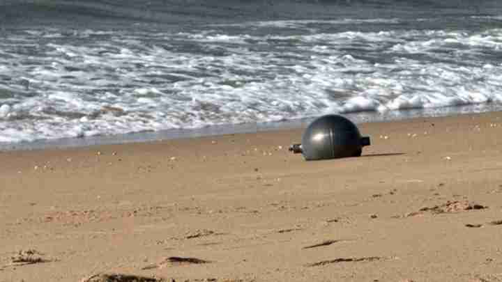 Black buoy washed up on a sandy beach near the ocean shoreline with small waves behind it.