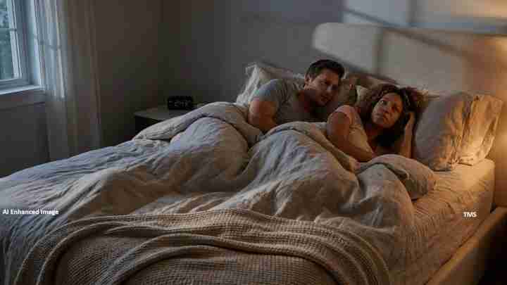 Couple lying in bed under a gray blanket in a dim bedroom, looking at the camera.