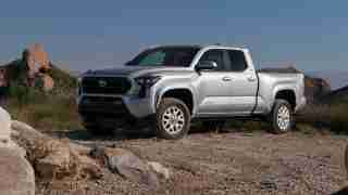 Front three-quarter view of a silver Toyota pickup parked on a rocky desert trail with boulders and scrubby plants under a clear blue sky.