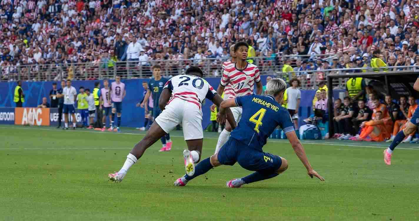Soccer players chase the ball as a blue defender slides in to tackle a white-red striped attacker, crowd watching from stands.