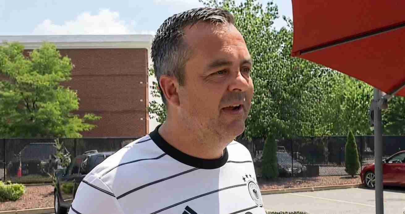 Middle-aged man with short gray hair wearing a white striped shirt speaks outdoors near a parking lot under a red umbrella.