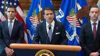 Three men in suits stand behind a podium with a Department of Justice seal, flanked by U.S. and DOJ flags.