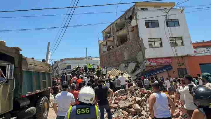 Crowd of rescuers and onlookers gather around a partially collapsed building, with rubble strewn across the street and a large truck on the left under a clear blue sky.