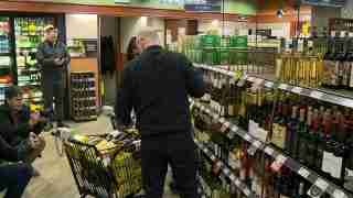 Candid shot in a liquor store: two men film with cameras while a man in black checks bottles in a cart.