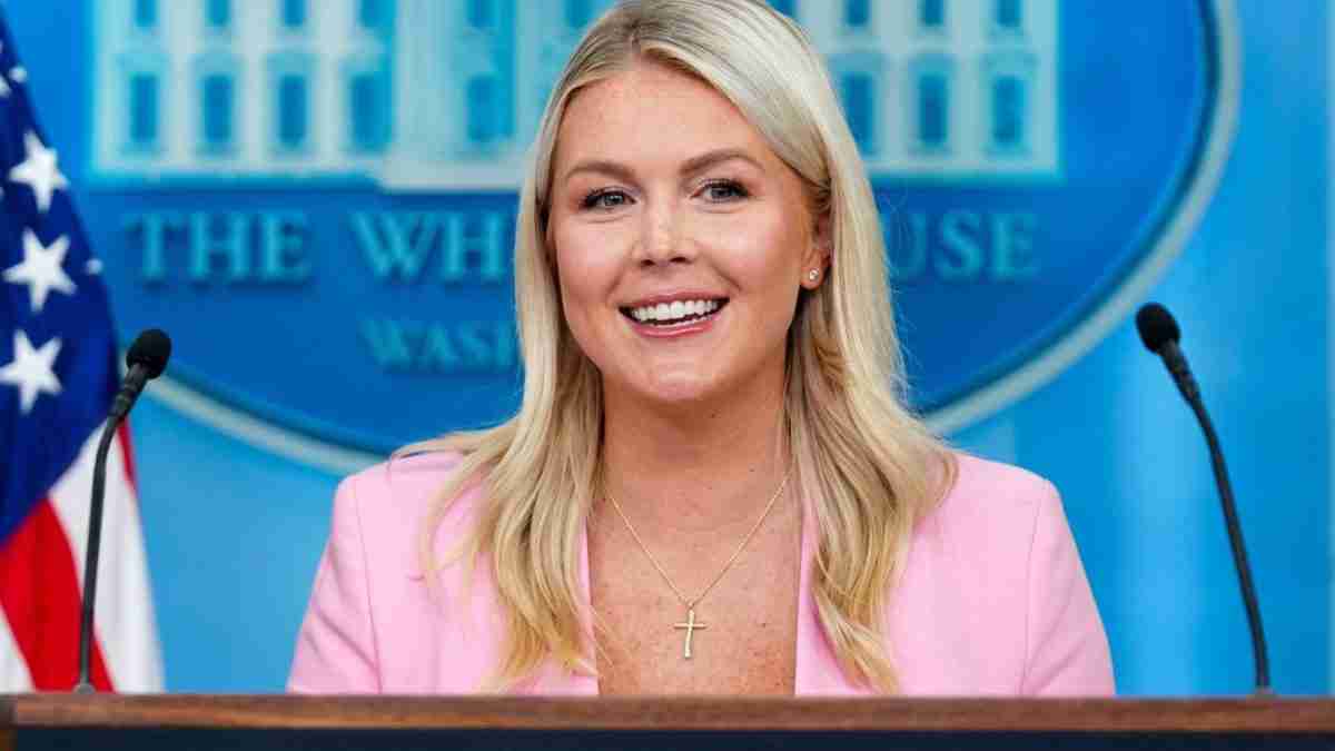 Speaker at a White House briefing wearing a pink blazer, smiling, with an American flag in the background and a cross necklace visible in front of a blue backdrop.