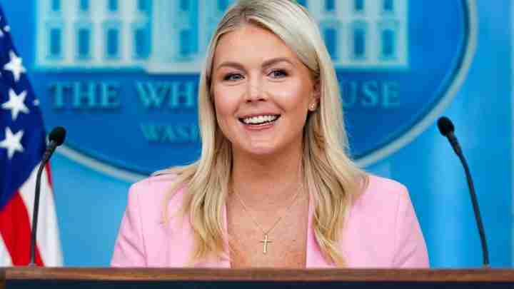 Speaker at a White House briefing wearing a pink blazer, smiling, with an American flag in the background and a cross necklace visible in front of a blue backdrop.