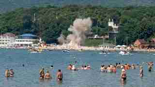Crowded lakeshore with people wading in water as a large smoky plume rises from the shore behind the beach buildings.