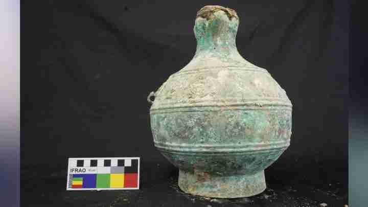 Ancient ceramic bottle with a narrow neck and bulbous body, green patina, placed on a black background beside a 10 cm measurement card.