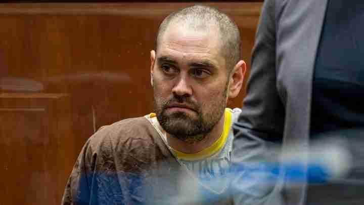 Defendant with short hair and beard sits in a courtroom wearing a brown jumpsuit, looking to the side.