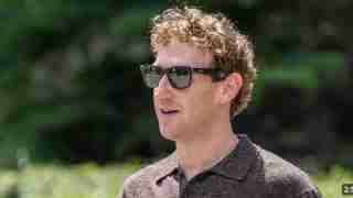 Man with curly blond hair wearing dark sunglasses outdoors, facing left.