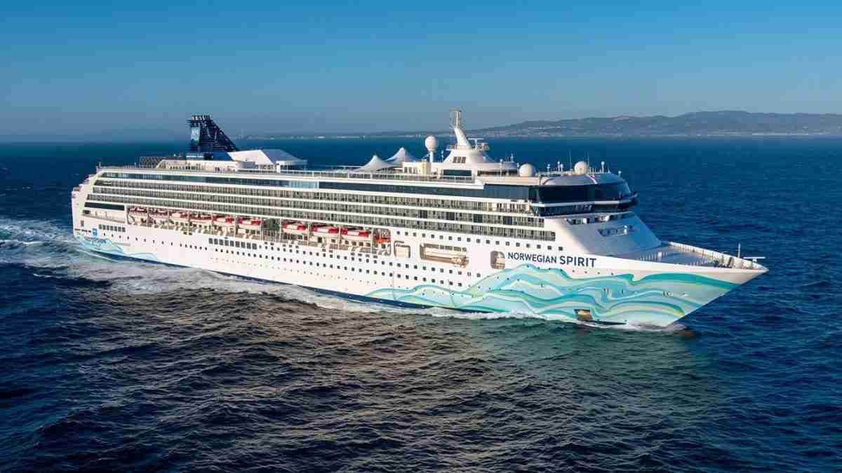 Large white cruise ship Norwegian Spirit sailing on calm blue sea with red-canopy decks and wave artwork on bow, coastline in distance.