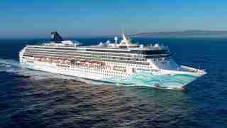 Large white cruise ship Norwegian Spirit sailing on calm blue sea with red-canopy decks and wave artwork on bow, coastline in distance.