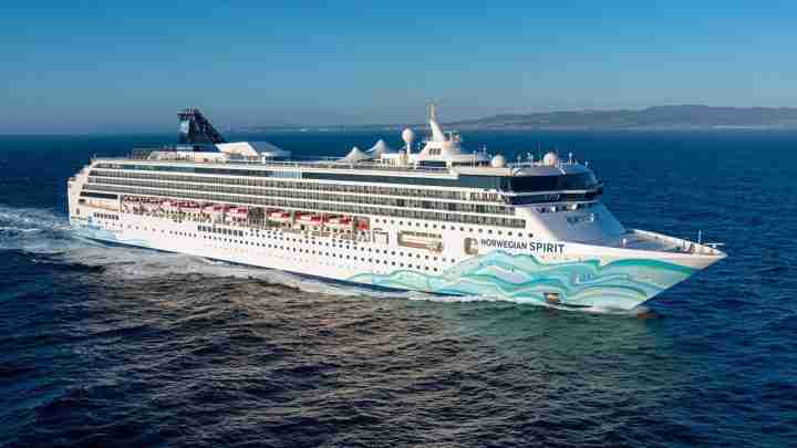 Large white cruise ship Norwegian Spirit sailing on calm blue sea with red-canopy decks and wave artwork on bow, coastline in distance.