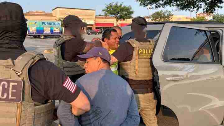 Police officers in tactical vests restrain a man beside a gray SUV in a parking lot.