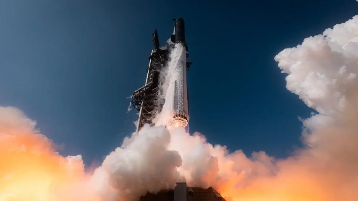 Rocket blasting off from a launch pad, with bright flame and white smoke at the base against a clear blue sky.
